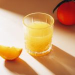 A glass of orange juice and slice of orange fruit on beige table with long hard shadows. Healthy raw vegan food, diet concept. Closeup.